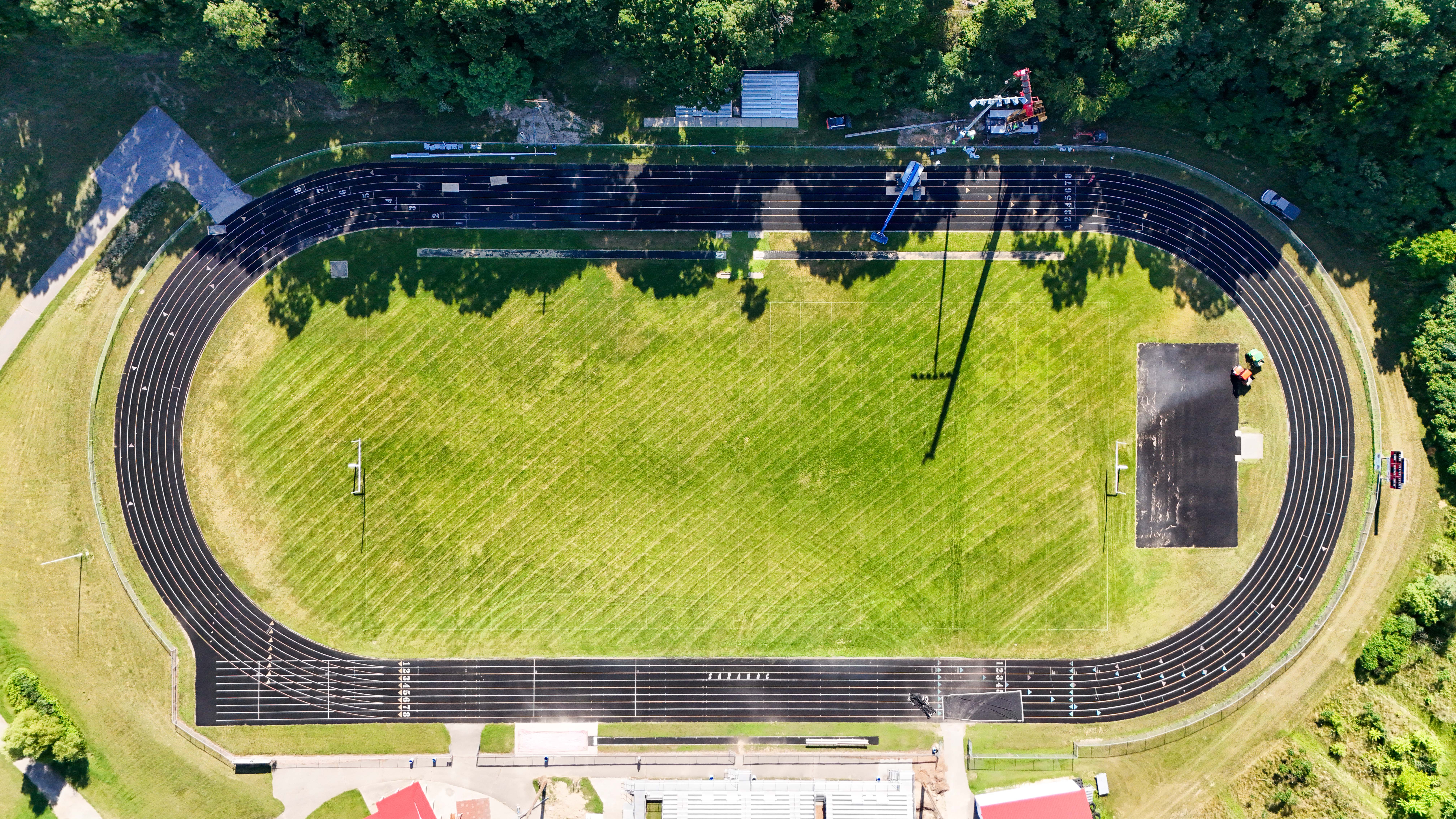 Aerial view of Saranac High School football field and track with Buist crew using a crane and lift to install new stadium lighting along the sideline.