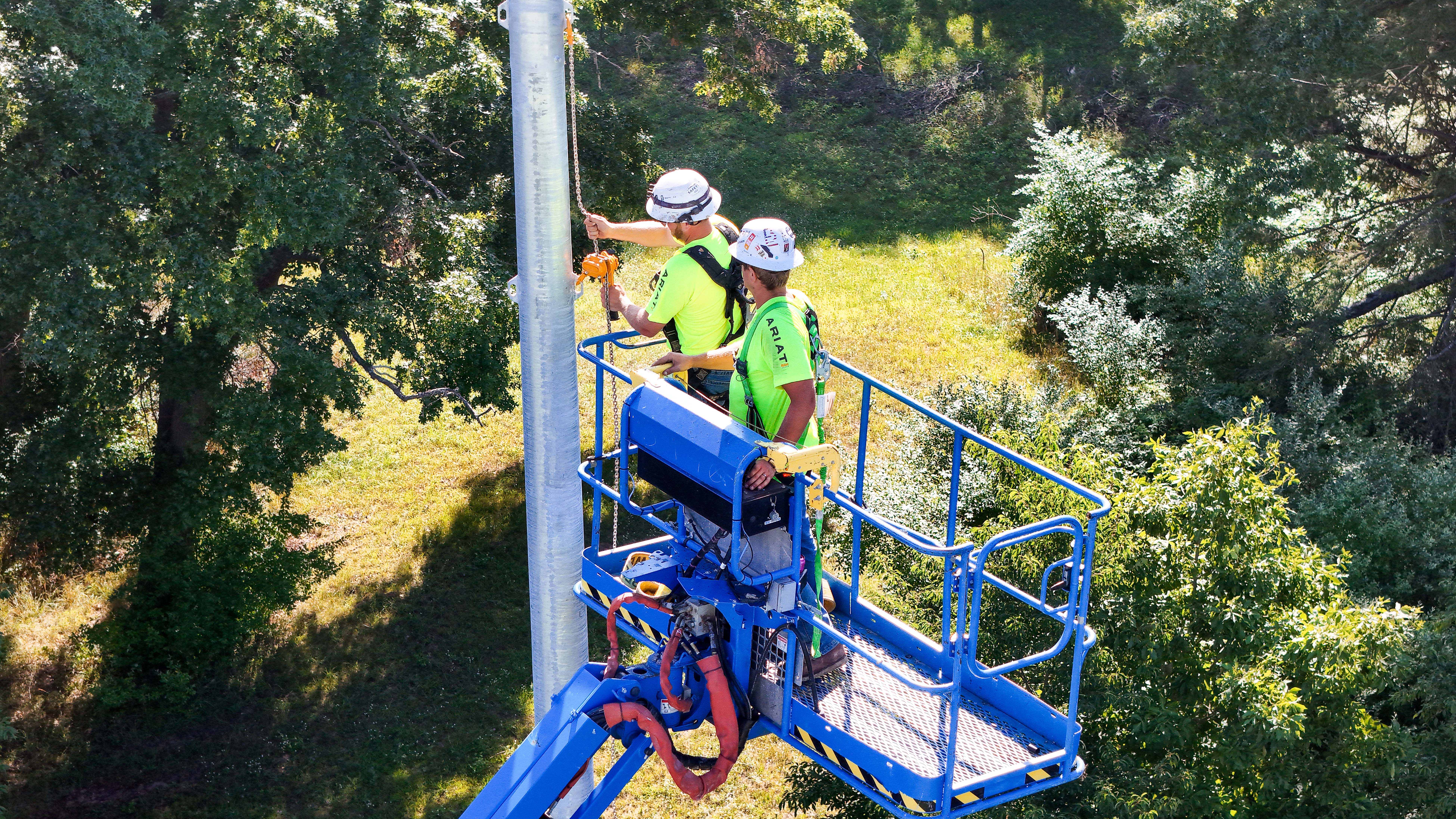 Two Buist electricians in safety gear working from an aerial lift to install components on a stadium light pole.