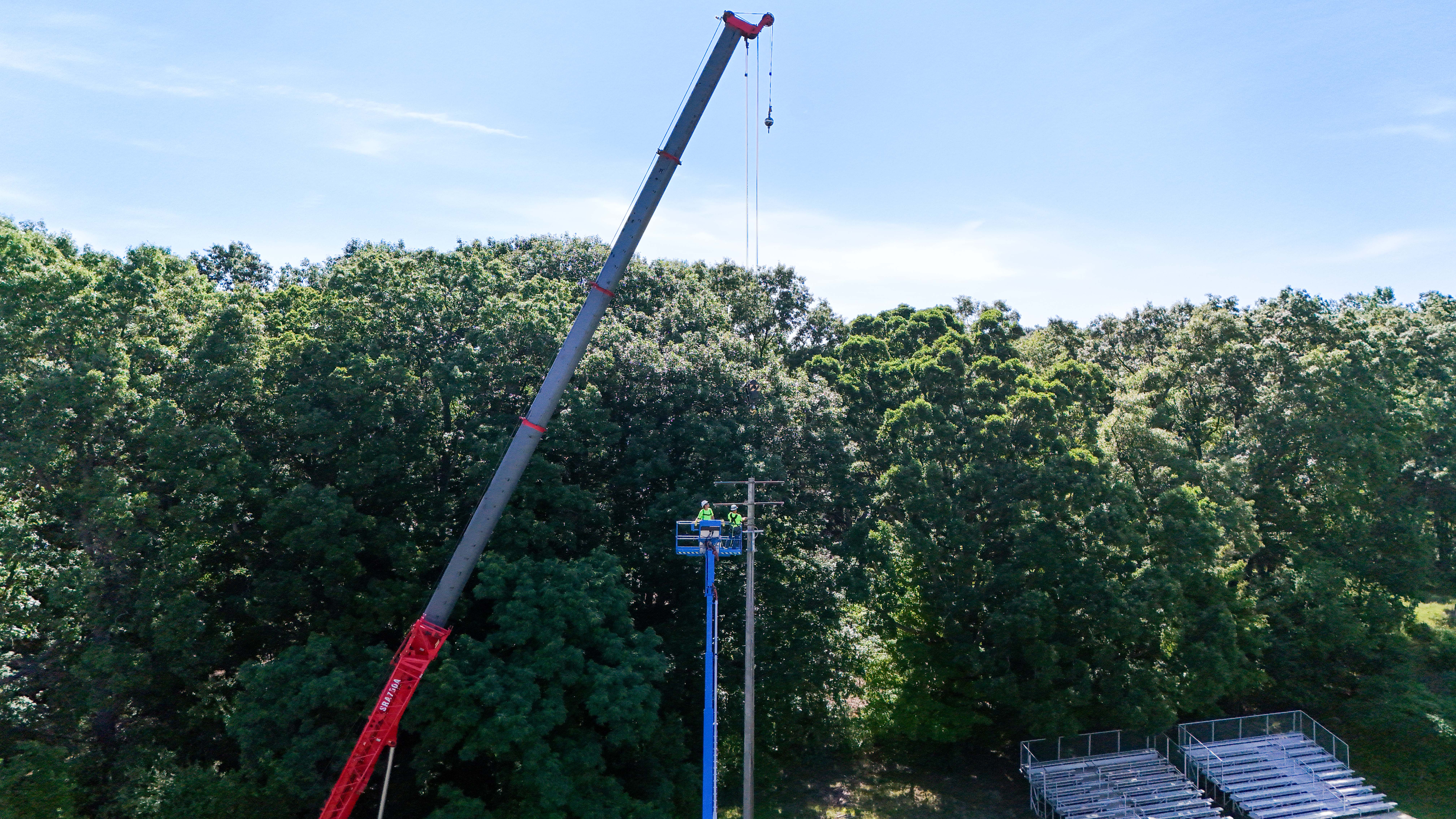Buist crew using a crane and aerial lift to install a stadium light pole at an Saranac High School surrounded by trees.