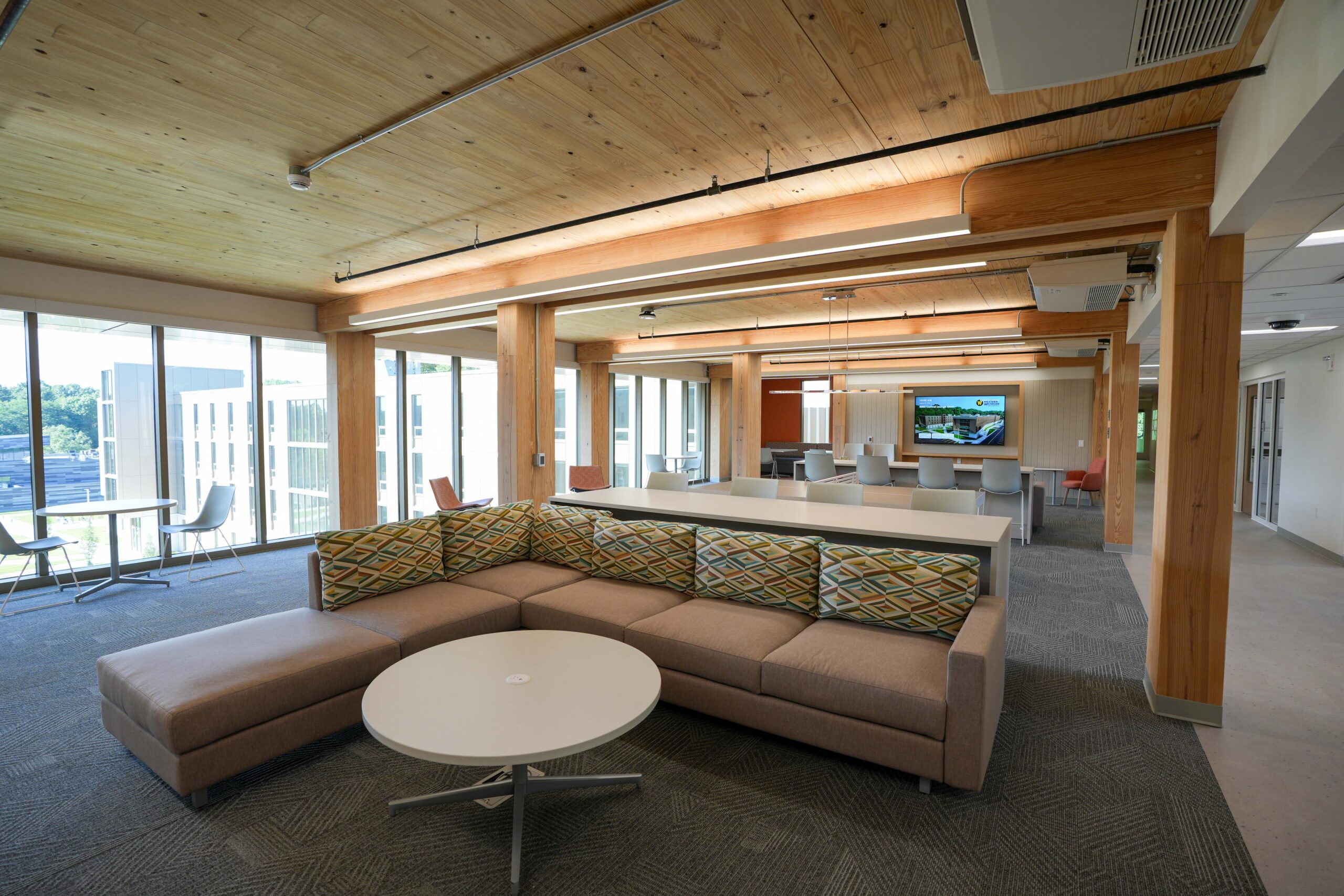 Western Michigan University Valley Oaks commons area with lounge seating, floor-to-ceiling windows, exposed timber columns, and a digital display.
