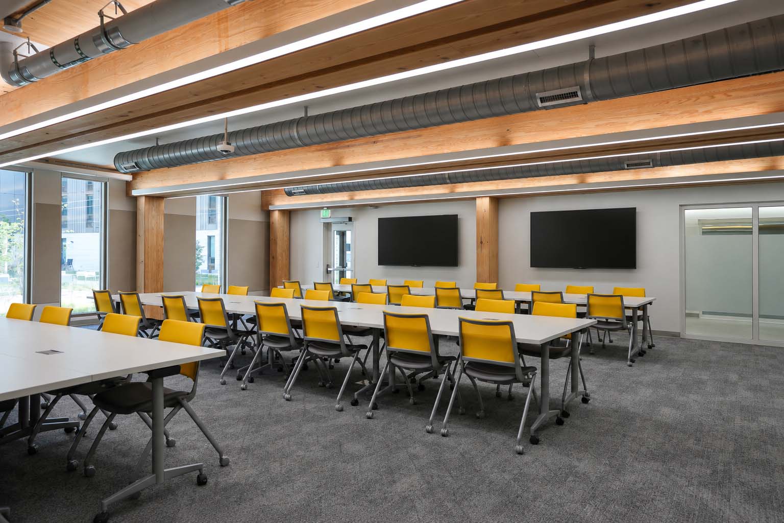WMU Valley Oaks multipurpose room featuring two wall-mounted commercial displays, movable tables, and yellow chairs.